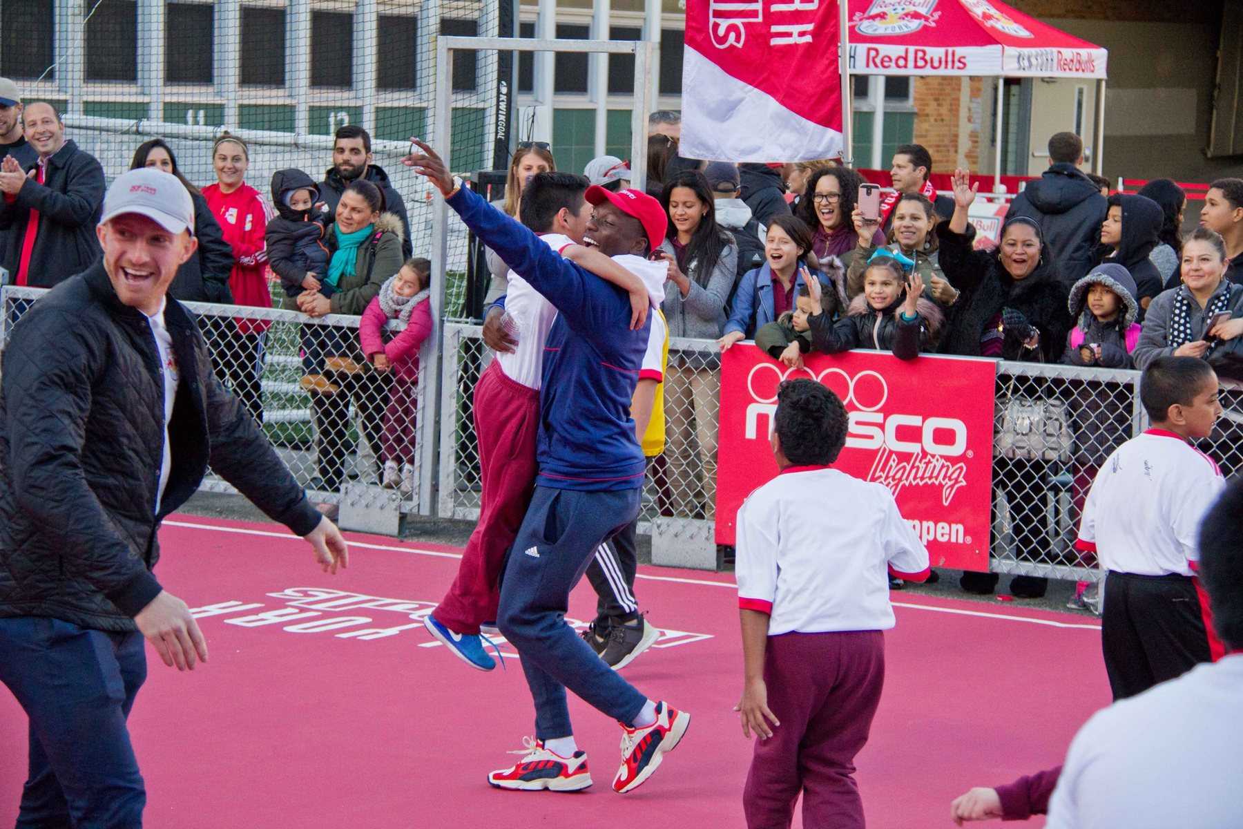 A coach lifts a young player as families celebrate around a neighborhood mini-pitch