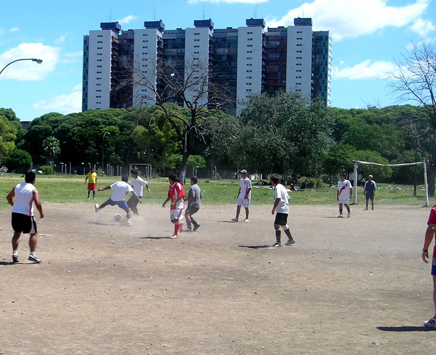 Players contesting the ball on a dirt potrero in Buenos Aires, with apartment blocks beyond the field